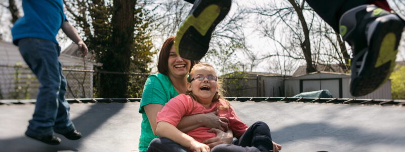 A woman and a girl sit laughing on a trampoline.