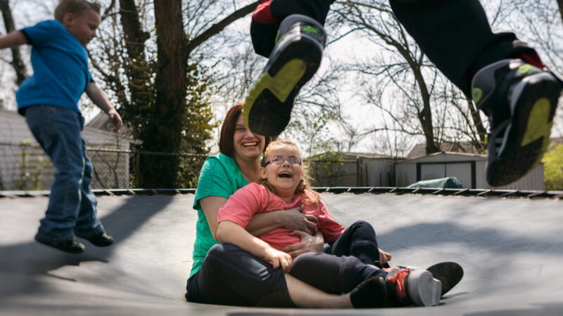 A woman and a girl sit laughing on a trampoline.